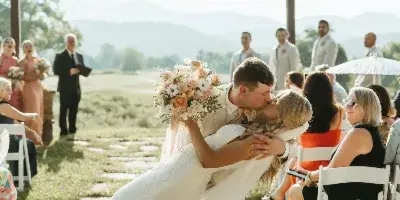 A bride and groom share a kiss during an outdoor wedding ceremony with an officiant and wedding party present, set against a backdrop of mountains.