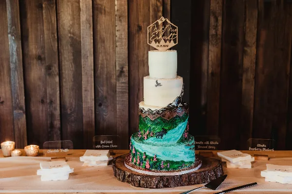 Four-tier wedding cake with white top tiers and a painted green mountain and turquoise waterfall design, topped with a hexagonal topper, set on a rustic wood slab with small flavor cards.