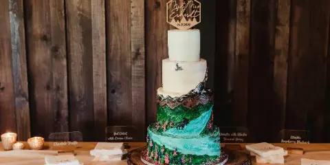 Four-tier wedding cake with white top tiers and a painted green mountain and turquoise waterfall design, topped with a hexagonal topper, set on a rustic wood slab with small flavor cards.