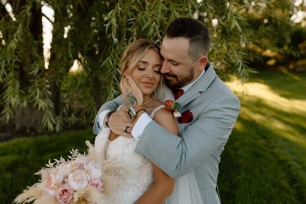 A bride and groom embracing outdoors with a bouquet of pink roses and pampas grass.