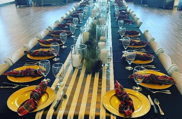 A long banquet table set for a formal event with gold plates, plaid napkins, glassware, and a centerpiece of candles and greenery.