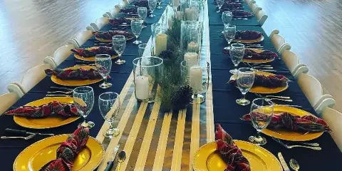 A long banquet table set for a formal event with gold plates, plaid napkins, glassware, and a centerpiece of candles and greenery.