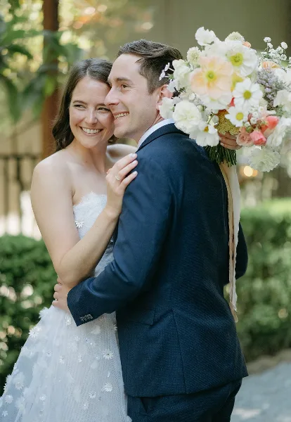 A bride and groom embracing outdoors, with the bride holding a large floral bouquet.