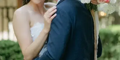 A bride and groom embracing outdoors, with the bride holding a large floral bouquet.