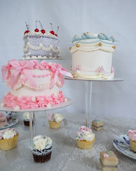 A display of three decorated cakes on stands surrounded by cupcakes and small pastries on a table.