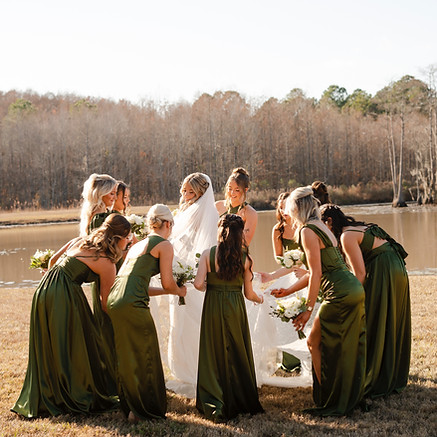 Bride and bridesmaids in green dresses posing outdoors near a pond with trees in the background.
