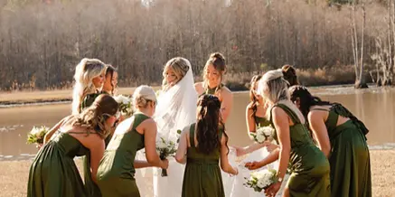 Bride and bridesmaids in green dresses posing outdoors near a pond with trees in the background.