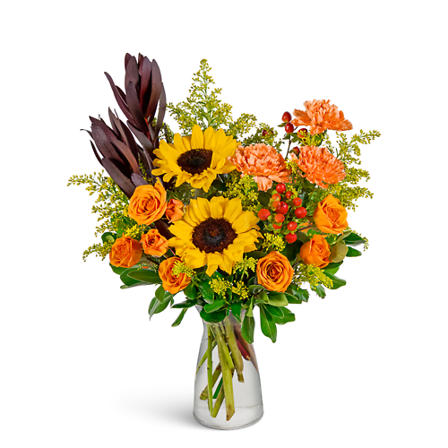 A floral arrangement featuring yellow sunflowers, orange roses, and greenery in a clear glass vase.