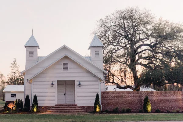 Exterior view of a white wooden chapel with twin spires and a brick wall at dusk.
