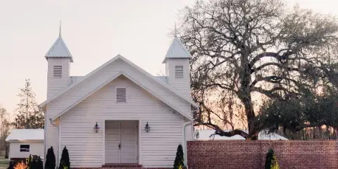 Exterior view of a white wooden chapel with twin spires and a brick wall at dusk.