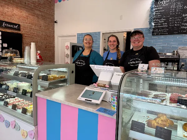Three staff members in Cake Me Away aprons standing behind a cafe counter with display cases of cakes and pastries.