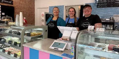 Three staff members in Cake Me Away aprons standing behind a cafe counter with display cases of cakes and pastries.