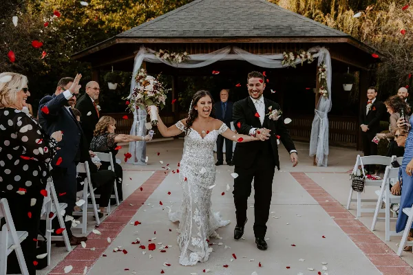A bride and groom walking down the aisle at an outdoor wedding ceremony with rose petals falling and guests seated on either side.