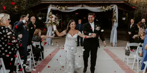 A bride and groom walking down the aisle at an outdoor wedding ceremony with rose petals falling and guests seated on either side.