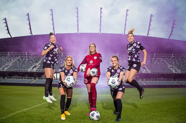 Five female soccer players in uniform posing with soccer balls on a stadium field.