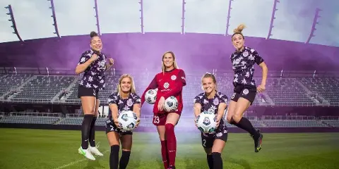 Five female soccer players in uniform posing with soccer balls on a stadium field.