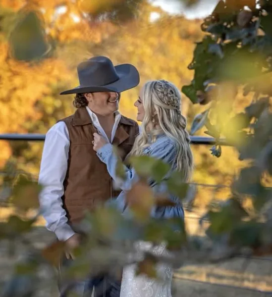 A bride and groom in wedding attire standing outdoors with autumn foliage in the background.