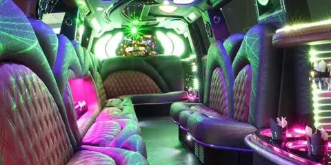 Interior view of a party bus featuring purple and green ambient lighting, plush leather seating, and a bar area.