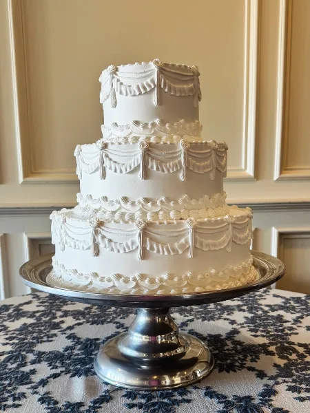 A three-tier white wedding cake with elaborate piped swag and floral details on a silver pedestal stand.