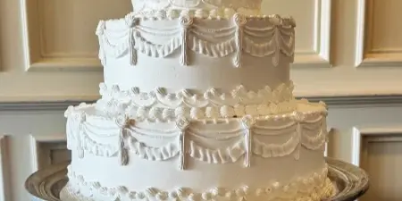 A three-tier white wedding cake with elaborate piped swag and floral details on a silver pedestal stand.