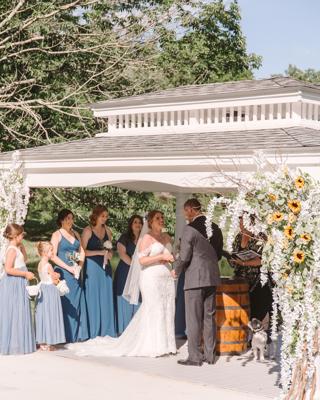 A bride and groom exchanging vows at an outdoor wedding ceremony under a rustic wooden pavilion decorated with white flowers and sunflowers.