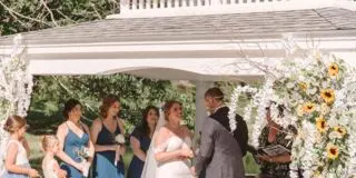 A bride and groom exchanging vows at an outdoor wedding ceremony under a rustic wooden pavilion decorated with white flowers and sunflowers.