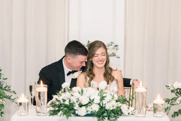 A bride and groom laughing together at a wedding reception table with floral centerpieces and candles.