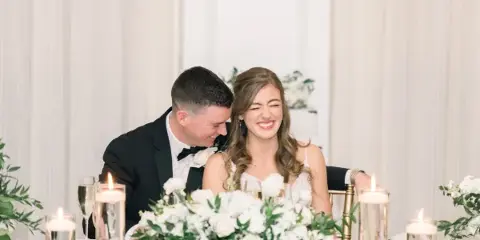 A bride and groom laughing together at a wedding reception table with floral centerpieces and candles.