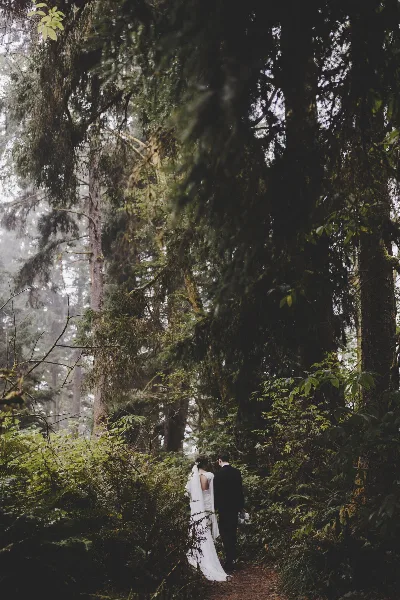 A bride and groom standing together on a path in a lush, green forest.