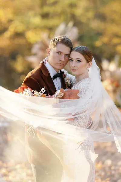 A bride and groom posing together in an outdoor setting with autumn foliage in the background.
