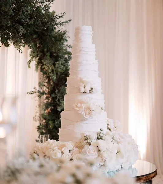 A tall, white, semi-naked wedding cake with multiple tiers, decorated with white roses and greenery, displayed on a stand in a bright, elegant setting.