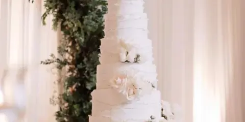 A tall, white, semi-naked wedding cake with multiple tiers, decorated with white roses and greenery, displayed on a stand in a bright, elegant setting.