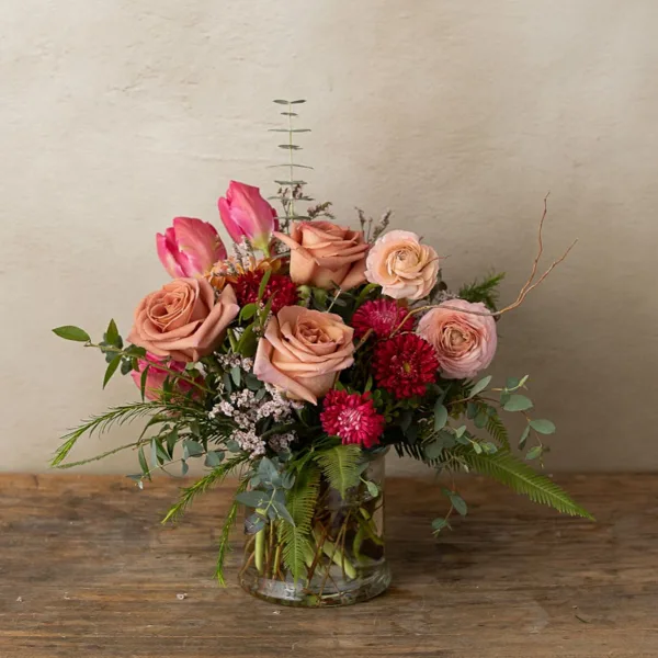 Fresh floral arrangement with peach roses, pink tulips, red dahlias, and greenery in a clear glass vase on a wooden table.