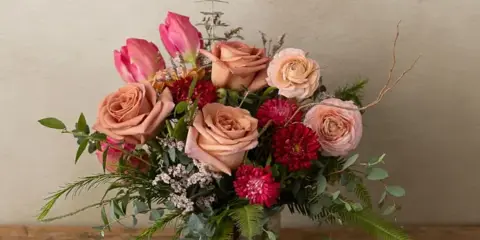 Fresh floral arrangement with peach roses, pink tulips, red dahlias, and greenery in a clear glass vase on a wooden table.