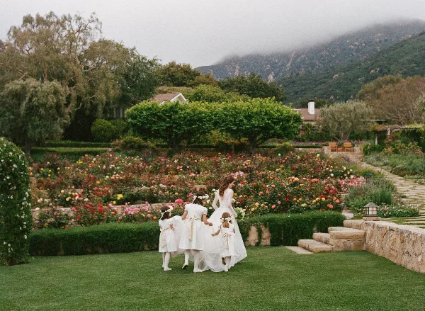 A bride and flower girls walking through a lush garden with a mountain backdrop.