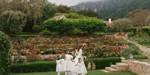 A bride and flower girls walking through a lush garden with a mountain backdrop.