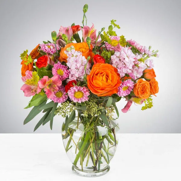 A vibrant mixed bouquet of orange, pink, and red flowers arranged in a clear glass vase.