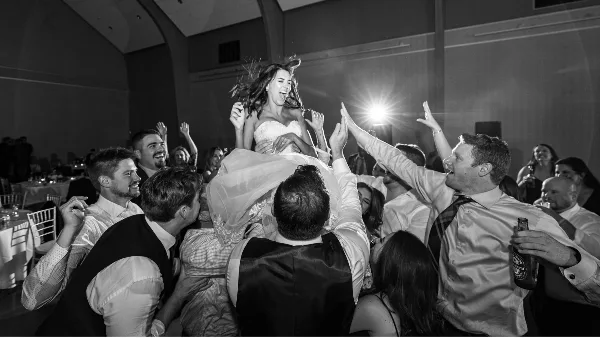 A black and white photo of a bride being lifted into the air by a group of cheering wedding guests during a reception.