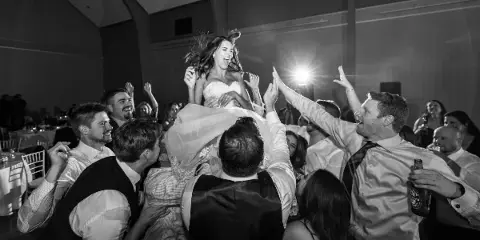 A black and white photo of a bride being lifted into the air by a group of cheering wedding guests during a reception.