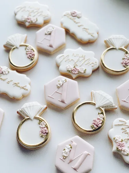 A close-up photograph of custom decorated sugar cookies featuring wedding ring shapes, monograms, and floral details in white, pink, and gold.