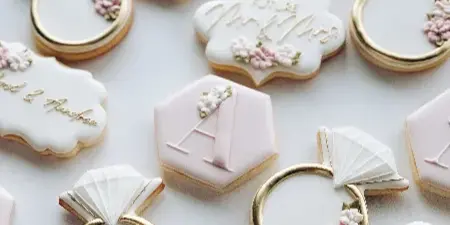 A close-up photograph of custom decorated sugar cookies featuring wedding ring shapes, monograms, and floral details in white, pink, and gold.