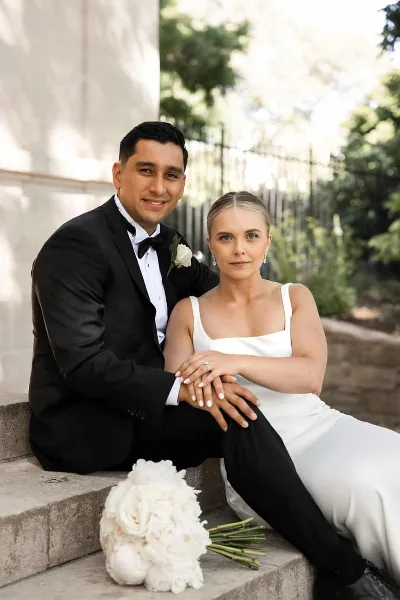 A bride and groom in formal wedding attire sitting on stone steps with a white bouquet.