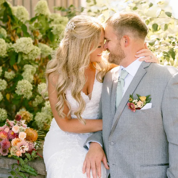 A bride and groom in wedding attire sharing an intimate moment outdoors with floral arrangements in the background.
