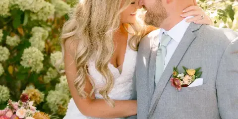 A bride and groom in wedding attire sharing an intimate moment outdoors with floral arrangements in the background.