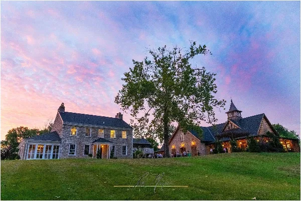 Exterior view of a stone wedding venue building with a large tree and lawn at dusk, with guests gathered near the entrance.