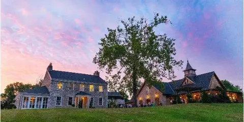 Exterior view of a stone wedding venue building with a large tree and lawn at dusk, with guests gathered near the entrance.