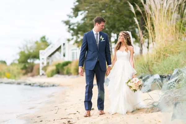 Bride and groom walking hand-in-hand on a sandy beach near water, with the bride holding a bouquet and a white venue structure in the background.