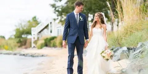 Bride and groom walking hand-in-hand on a sandy beach near water, with the bride holding a bouquet and a white venue structure in the background.