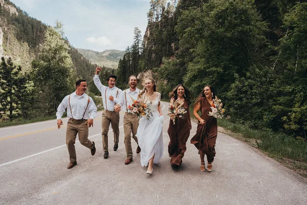 A wedding party consisting of a bride, groom, and four attendants running joyfully down a paved road surrounded by forested hills.