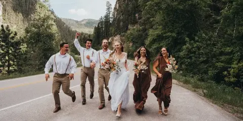 A wedding party consisting of a bride, groom, and four attendants running joyfully down a paved road surrounded by forested hills.